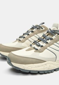 Sneakers mają beżowo-szary kolor, z gładką tekstylną cholewką, zamszowymi akcentami, plamkowanym śródsołem i szarymi sznurowadłami.