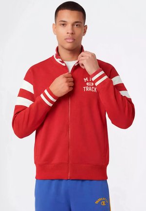 Jonge man die een rood trainingsjack met witte strepen op de mouwen dichtritst, en blauwe broek met gele merkopdruk draagt.