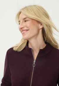 Bordeauxrode trui met rits en hoge kraag, gestructureerde stof en zilveren rits met een ronde trekker. Model heeft lang blond haar.