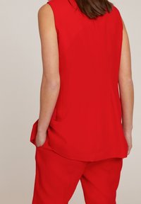 Haut sans manches rouge avec une texture lisse et des poches latérales, présentant un ajustement légèrement ample et des lignes épurées, associé à un pantalon assorti.