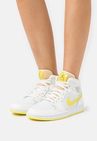 Person wearing white and yellow high-top Nike sneakers with laces, standing against a plain white background.
