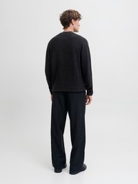 Mladý muž s kučeravými vlasmi stojí chrbtom, oblečený v čiernom svetri, nohaviciach s širokými nohavicami s prúžkami a čiernych topánkach na obyčajnom pozadí.