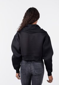 Sudadera negra recortada con un cuello alto y relajado y mangas anchas, con una textura suave y puños acanalados. Combinada con jeans oscuros.
