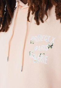 Sudadera color durazno con el texto blanco "PROJECT X PARIS GALERIE" rodeado de acentos florales; cuenta con cordones con puntas metálicas; textura de mezcla de algodón.