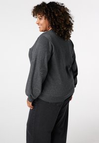 Glunderende vrouw met krullend haar, gekleed in een donkergrijze trui met hartpatronen op de mouwen en zwarte broek, staande tegen een witte achtergrond.