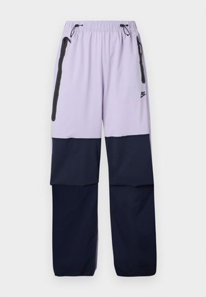 Fialové a námornícke nohavice Nike s elastickým pásom, šnúrkou, bočnými zipsovými vreckami a farebným blokovým dizajnom. Vyrobené z ľahkej tkaniny.