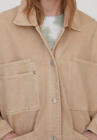 Veste en denim beige avec col pointu, deux poches avant et boutons-pression argentés, présentant une texture douce.
