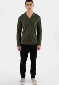 Langarm-Polohemd aus dunkelgrünem Baumwollmaterial mit V-Ausschnitt, kombiniert mit schwarzen Hosen und beigen Sneakers. Fitted Design und glatte Textur.