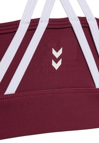 Zbliżenie bordowego biustonosza sportowego z białym podwójnym logo w kształcie jodełki i białymi przeszywanymi ramiączkami krzyżującymi się z tyłu.