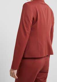 Blazer rouge ajusté avec une coupe cintrée, présentant un col cranté et deux poches latérales, confectionné dans un tissu texturé.