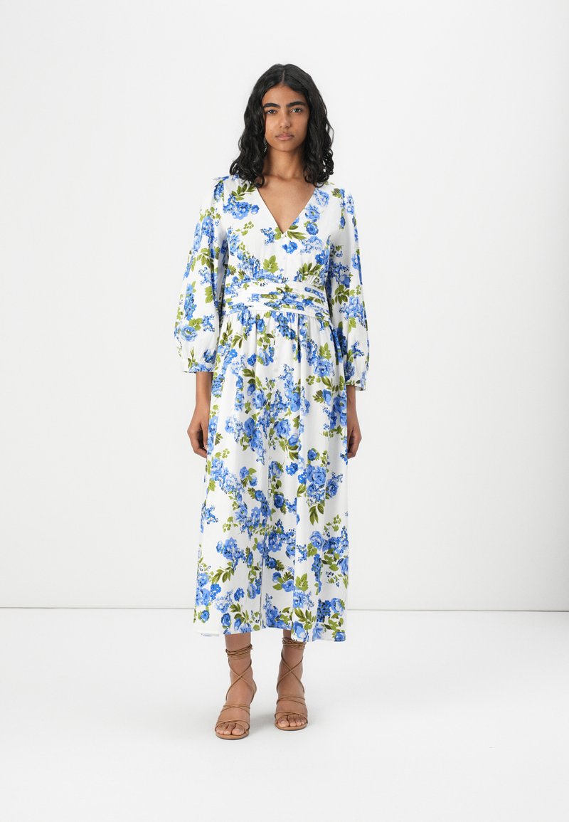 Robe midi florale avec une base blanche ornée de motifs floraux bleus et verts, décolleté en V profond, manches longues et ceinture à la taille. Complétée par des sandales à lanières marron.