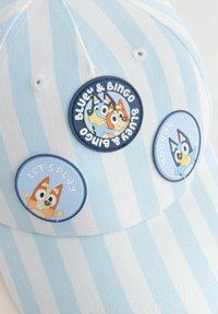 Gorra a rayas azul y blanca con tres pines circulares: dos con los personajes "Bluey y Bingo" y uno que dice "Vamos a jugar". Textura de tela suave.