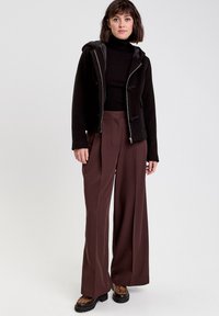 Sort zip-up jakke med hætte, parret med en tætsiddende sort rullekrave og brede brune bukser. Kraftige sorte sko med leopardmønster accenter.