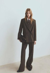 Completo doppiopetto a quadri marroni con pantaloni a gamba larga, abbinato a una camicia bianca e una cravatta scura. Il modello tiene in mano una pochette nera.