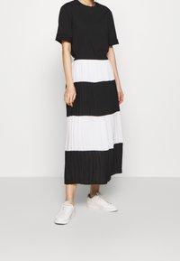 Schwarzer und weißer gefalteter Rock mit breiten horizontalen Streifen, kombiniert mit einem schwarzen Kurzarmshirt und weißen Turnschuhen. Glatte Stofftextur.