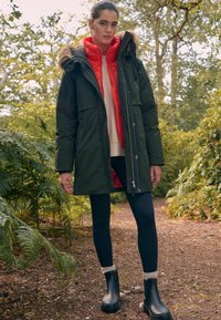 Parka verte avec fourrure, superposée à une doudoune rouge vif. Leggings noirs et bottines à cheville avec accents blancs. Entourée de verdure.