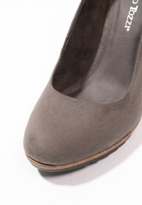 Chaussure à talon haut en daim gris avec un bout arrondi, des détails cousus et une légère plateforme. Elle présente une texture lisse et une semelle en caoutchouc.