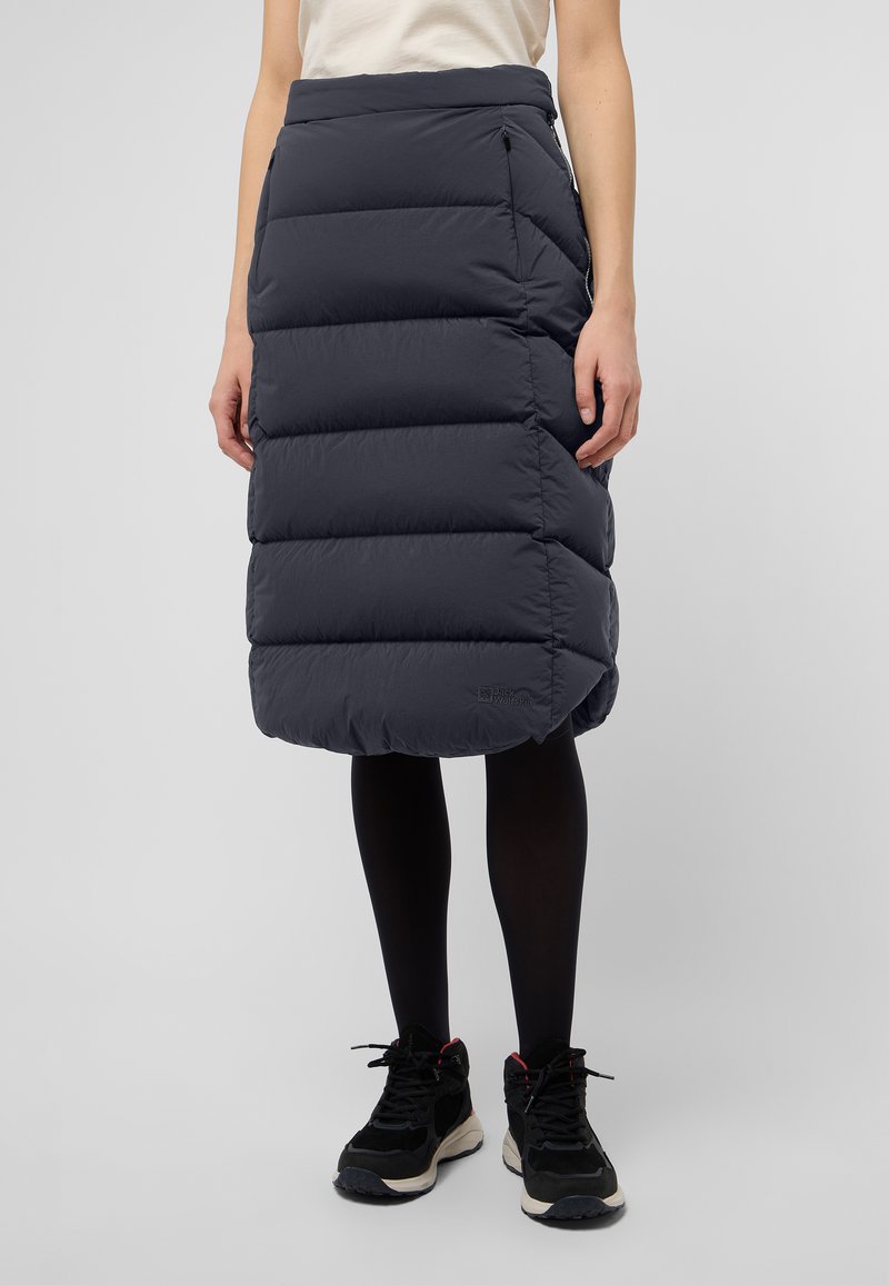 Jupe isolante grise et rembourrée avec une taille haute, des poches latérales zippées et un ourlet au genou. Portée avec des leggings noirs et des baskets.