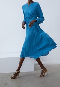 En blå plisserad midiklänning med nyckelhålsringning och bälte i midjan, tillsammans med guldiga remsandaler med hög klack. Slät yta.