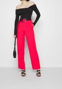 Svart off-the-shoulder topp med långärmar, i kombination med höga rosa vida byxor. Svarta sandaletter med remmar och en svart handväska.