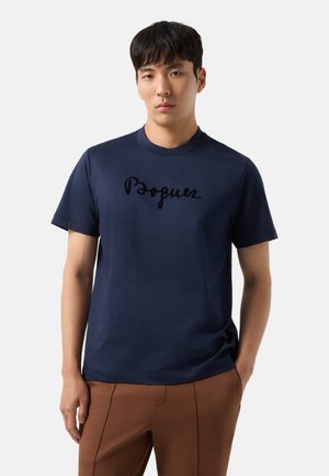 Ung mand med kort sort hår iført en navyblå t-shirt med sort skrifttekst og brune bukser, står med den ene hånd i lommen.