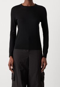 Personne portant un t-shirt noir ajusté à manches longues et un pantalon cargo noir ample avec des poches, debout devant un fond uni.