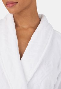 Biały bawełniany szlafrok o kroju V-neck, miękka tekstura i haftowany detal z logo. Zbliżenie z widokiem na kołnierz szalowy.