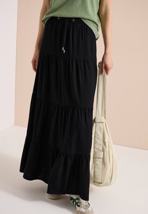 Osoba nosząca czarną, wielowarstwową maxi spódnicę, zielony top, zielono-białe trampki i trzymająca beżową torebkę na ramię przy boku.