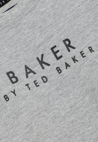 Pelēkas kokvilnas T-kreklas ar teksturētu uzdruku, kurā redzams vārds "BAKER" treknā melnā burtu un "by TED BAKER" mazākā tekstā zem tā.