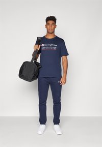 Navy Champion athletic t-shirt with white and red logo, matching navy sweatpants, and black duffel bag, held with a single strap. White sneakers.