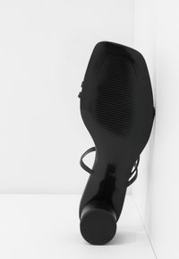 Chaussure à talons hauts noire avec un bout arrondi, des fines lanières et une semelle texturée. Taille indiquée comme 38. Design épuré et minimaliste sur un fond blanc.