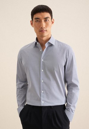 Camicia abbottonata di colore azzurro chiaro con motivo geometrico. Maniche lunghe, colletto alla francese e tessuto liscio e strutturato. Abbinata a pantaloni scuri.