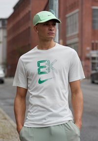 Hvit Nike t-skjorte med grønt grafisk design og logo, kombinert med lysegrønne shorts. Modell iført en lys grønn caps. Urban bakgrunn.