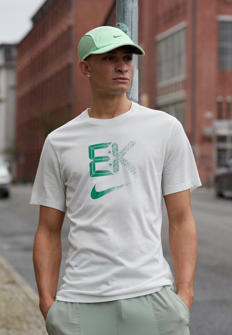 Hvit Nike t-skjorte med grønt grafisk design og logo, kombinert med lysegrønne shorts. Modell iført en lys grønn caps. Urban bakgrunn.