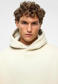 Beige hoodie met een grote, dubbele capuchon. Glad textuur van de stof, accentueert met twee zilveren drukkers bij de nek. Minimalistisch ontwerp.