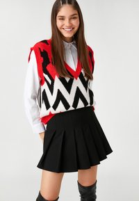 Czerwono-czarny sweter w deseń z dekoltem w serek, nałożony na biały shirt; zestawiony z czarną spódnicą plisowaną. Gładkie tekstury.
