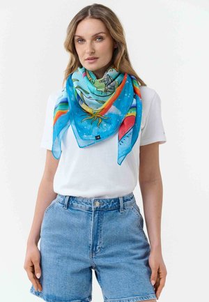 Woman wearing blue patterned scarf with rainbow and nature motifs, white t-shirt, and high-waisted denim shorts, standing against white background.