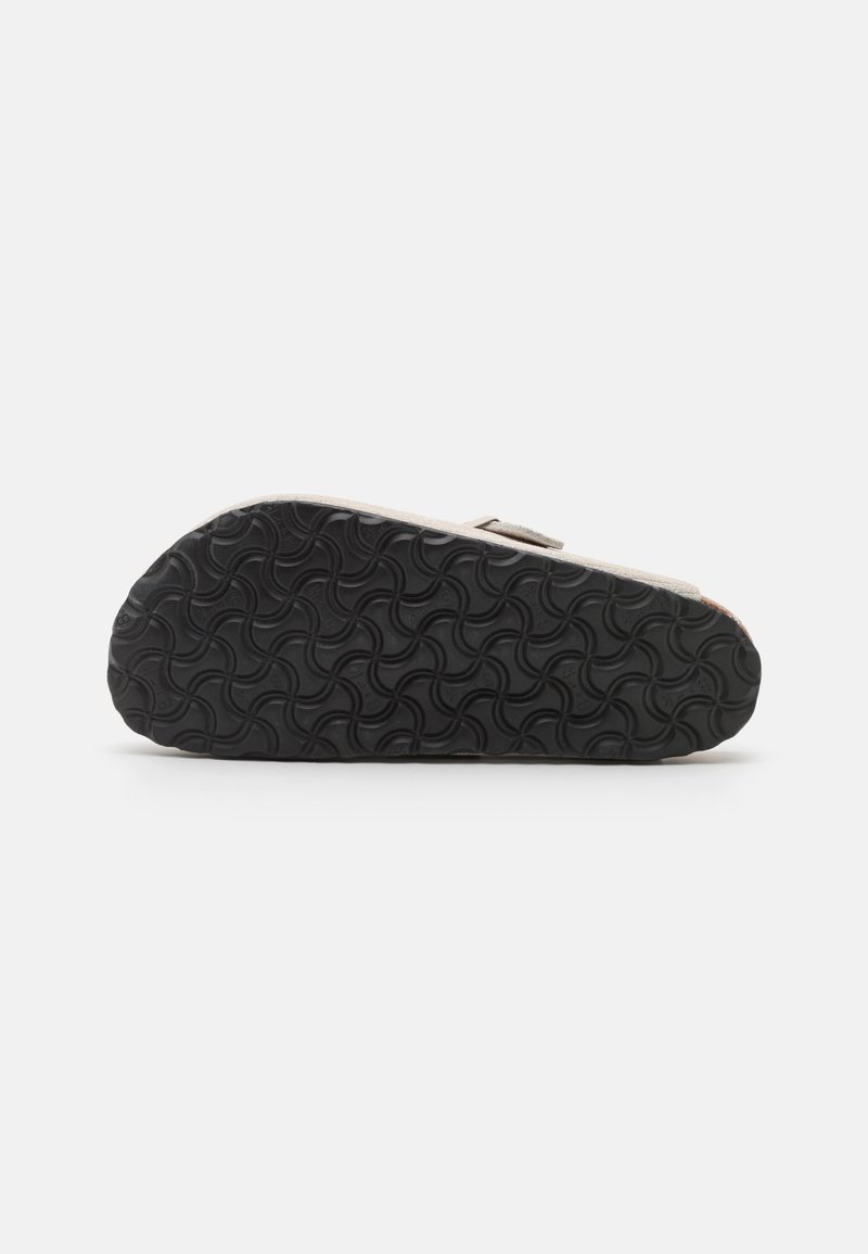 La suela del zapato cuenta con un caucho negro texturizado con un patrón ondulado; la parte superior muestra un acabado de ante beige claro y un diseño de tira simple.
