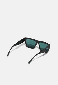 Ochelari de soare negri cu rame unghiulare, lentile de culoare teal și un logo "CK" albastru pe lateral. Textură plastică mată, design elegant.