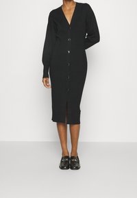 Robe cardigan noire en maille avec un col en V, des manches longues, une fermeture boutonnée à l'avant et une fente centrale à l'ourlet. Texture lisse, coupe ajustée.