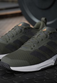 Paire de chaussures de sport vertes et noires avec des semelles blanches placées sur un sol noir texturé près du matériel d'exercice.