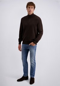 Uomo che indossa un pullover marrone scuro con zip al quarto, jeans azzurri chiari e stivali marrone scuro, in piedi contro uno sfondo grigio uniforme.