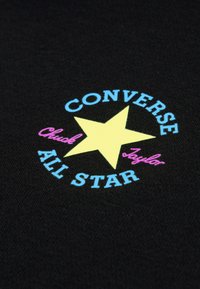 Μαύρο ύφασμα με στρογγυλό λογότυπο που έχει ένα κίτρινο αστέρι. Το κείμενο περιλαμβάνει το "CONVERSE" σε μπλε, το "Chuck" σε ροζ και το "ALL STAR" σε μπλε.