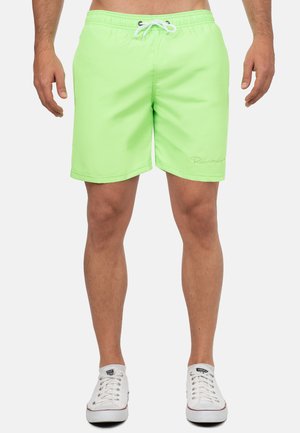 Man die felgroene zwembroek met een wit trekkoord en witte sneakers draagt, staand tegen een effen achtergrond.