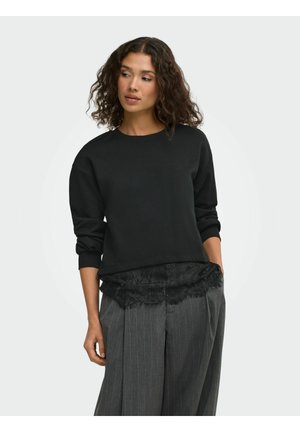 Femme aux cheveux bouclés portant un haut noir à manches longues avec un détail en dentelle et un pantalon large gris à fines rayures, les mains dans les poches.