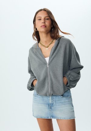 Siva zip-up dukserica s ležernim krojem i elastičnim manžetama, uparena s svijetloplavom traper mini suknjom. Model nosi ogrlice u više slojeva.