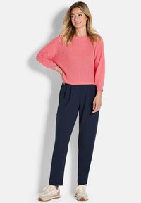 Vrouw in een koraalrode ribgebreide trui met lange mouwen, marineblauwe plooibroek en beige sneakers, staand met één hand op haar heup.