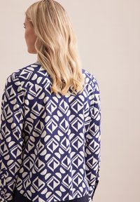 Camicia a maniche lunghe in tessuto blu navy con motivo geometrico bianco. Presenta una spalla posteriore e bottoni ai polsini, texture liscia.
