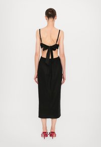 Zwart midi-jurk met een strikdetail op de rug, dunne bandjes en een aansluitende taille. Model draagt roze hoge hakken. Gladde stoftextuur.
