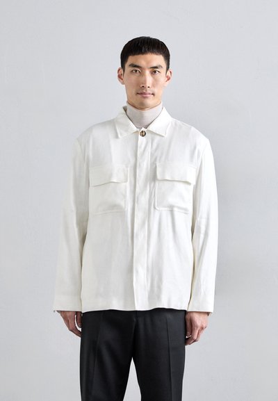 Veste blanche à manches longues en tissu texturé ; dotée d'un col, de deux poches poitrine et d'une fermeture à boutons. Portée par-dessus un col roulé léger.